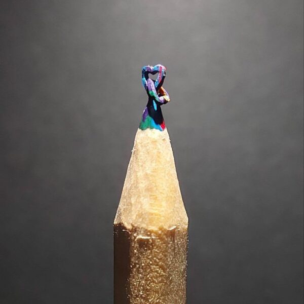 Close-up of two intricately carved hearts entangled together at the tip of a sharpened pencil, showcasing detailed miniature craftsmanship and delicate design on graphite.