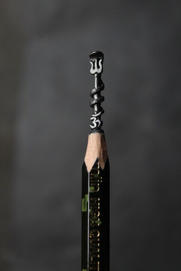 Close-up of a hand-carved Trishul placed above an Om symbol on the graphite tip of a pencil, symbolizing Lord Shiva and spiritual craftsmanship in miniature form