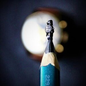Macro image of a detailed miniature watch sculpture carved into the graphite tip of a pencil, showcasing tiny dial and strap details with fine craftsmanship
